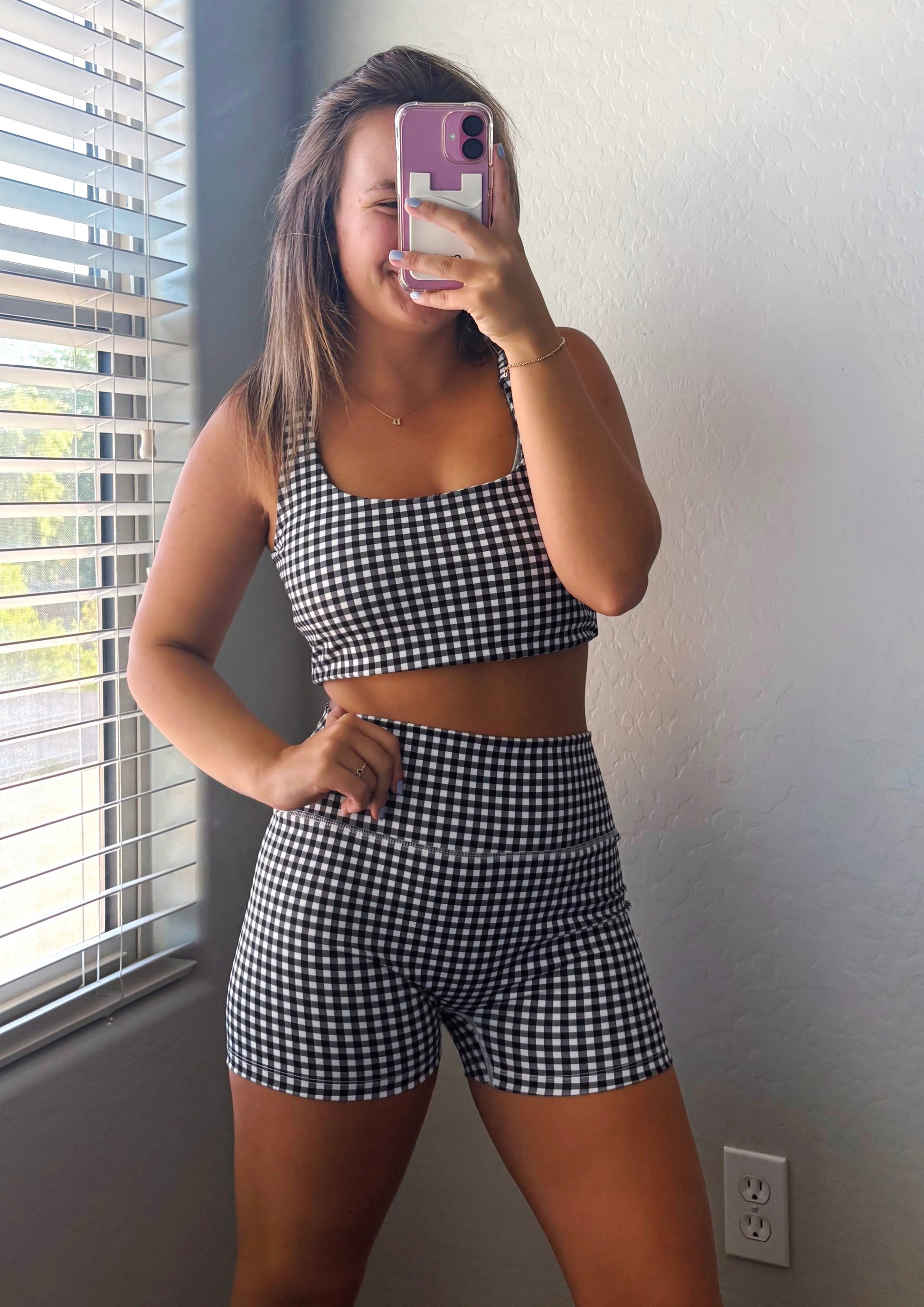 Person wearing a checkered outfit taking a mirror selfie in a room with a window and bed.